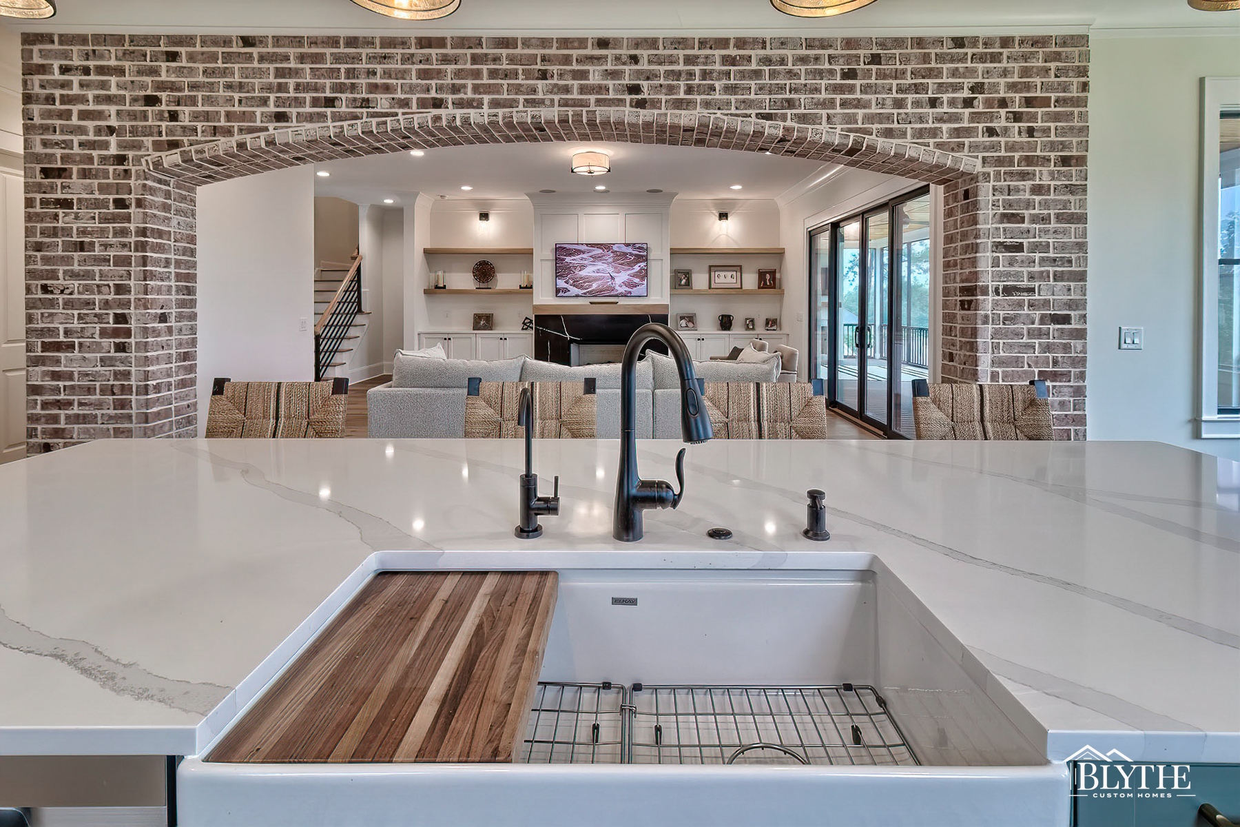 White farmhouse sink with wood cutting board insert, antique bronze-look faucet, and white quartz counter with light gray veining and large brick archway leading to the living space with fireplace and built-ins.