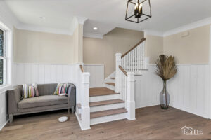 The foyer with love seat and L-shaped wooden stairs and white wainscoting with rustic hardwood floors.