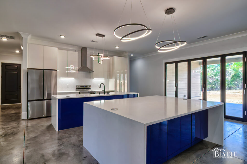 An industrial-style kitchen with polished concrete floors and two large white kitchen islands with deep royal blue cabinets and modern LED pendant lights hanging over the eat-in island.