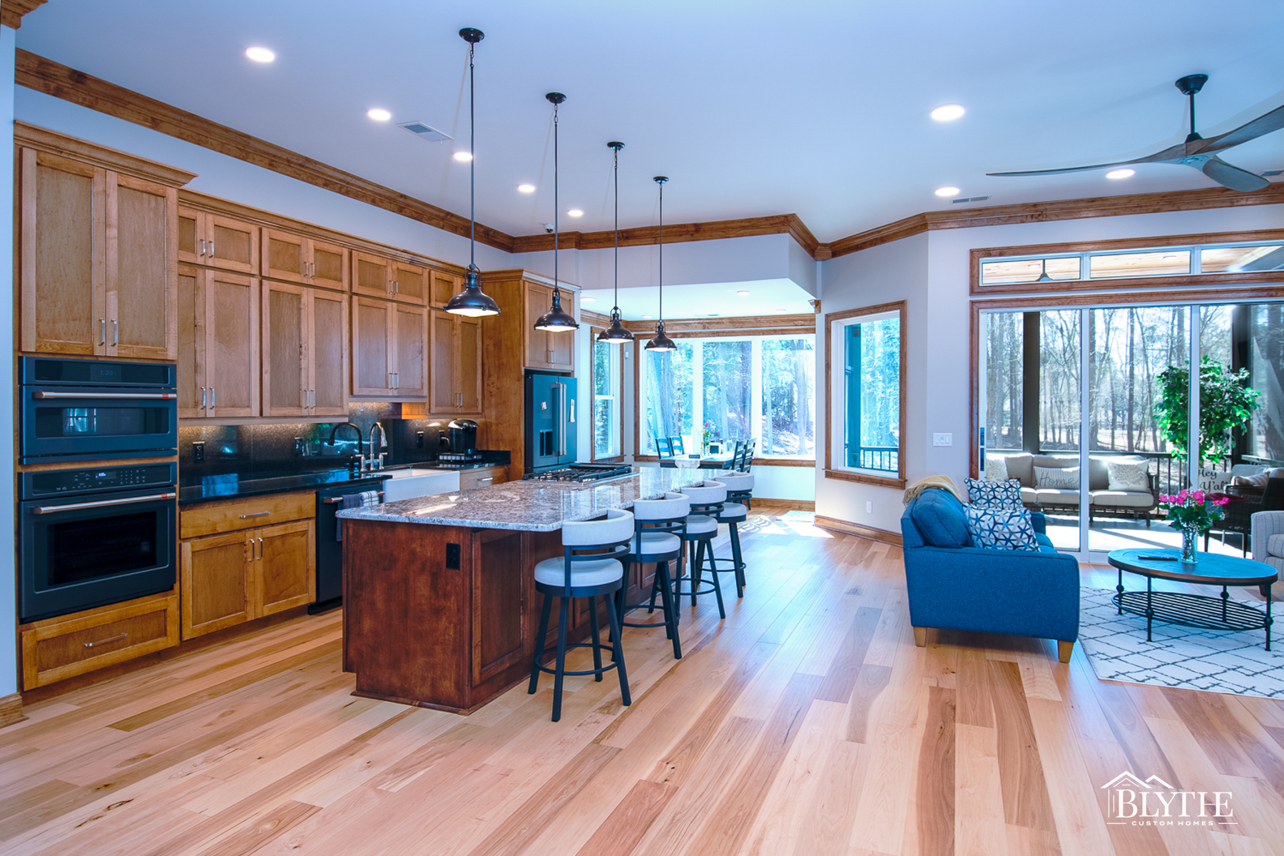 Stained solid wood kitchen cabinets with black appliances, dark stained wood island with 4 pendant lights, natural hickory hardwood floors, and a wall full of windows facing the woods.