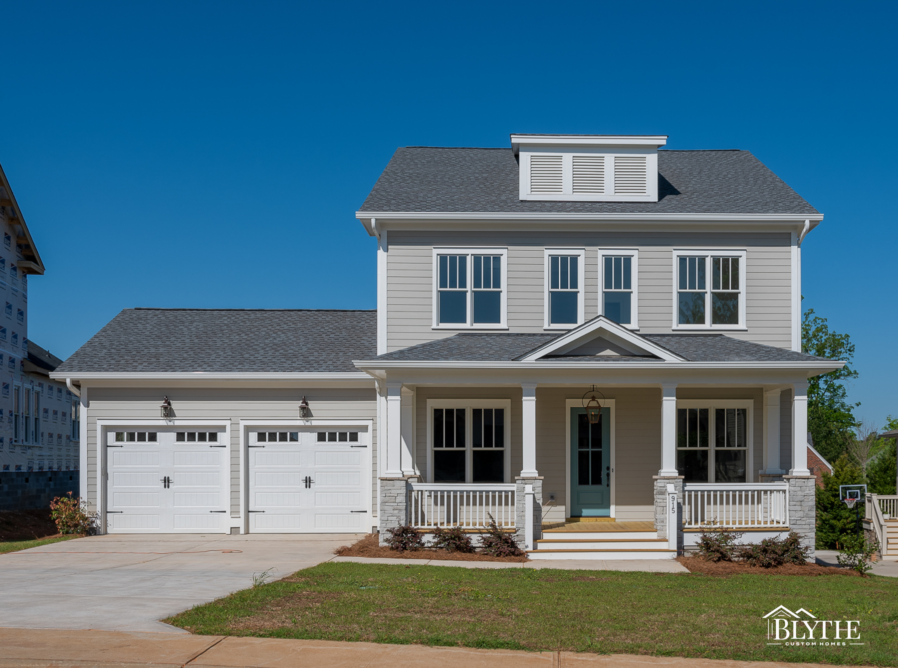 2-story home with gray Hardie plank siding, a front porch with white columns and stone bases, and 2-car garage with single carriage garage doors on sunny day with blue sky.
