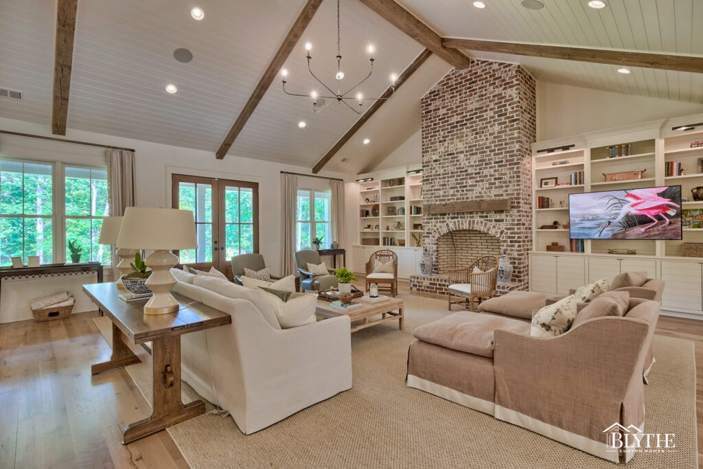 A huge living space with vaulted wood-covered ceiling with the wood painted white and rustic wood ceiling beams and a huge masonry fireplace with brick that reaches to the tall ceiling and built-ins on each side.