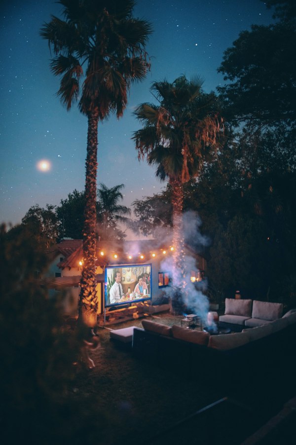 Patio at night with patio string lights, fire pit, and movie screen and palm trees and moon and stars