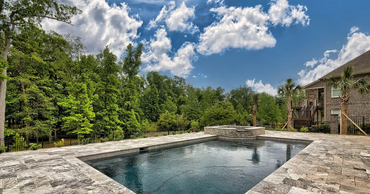 A custom pool with silver travertine pool deck and pool coping and a custom spa with a view of the woods behind in spring on a partly cloudy and sunny day.