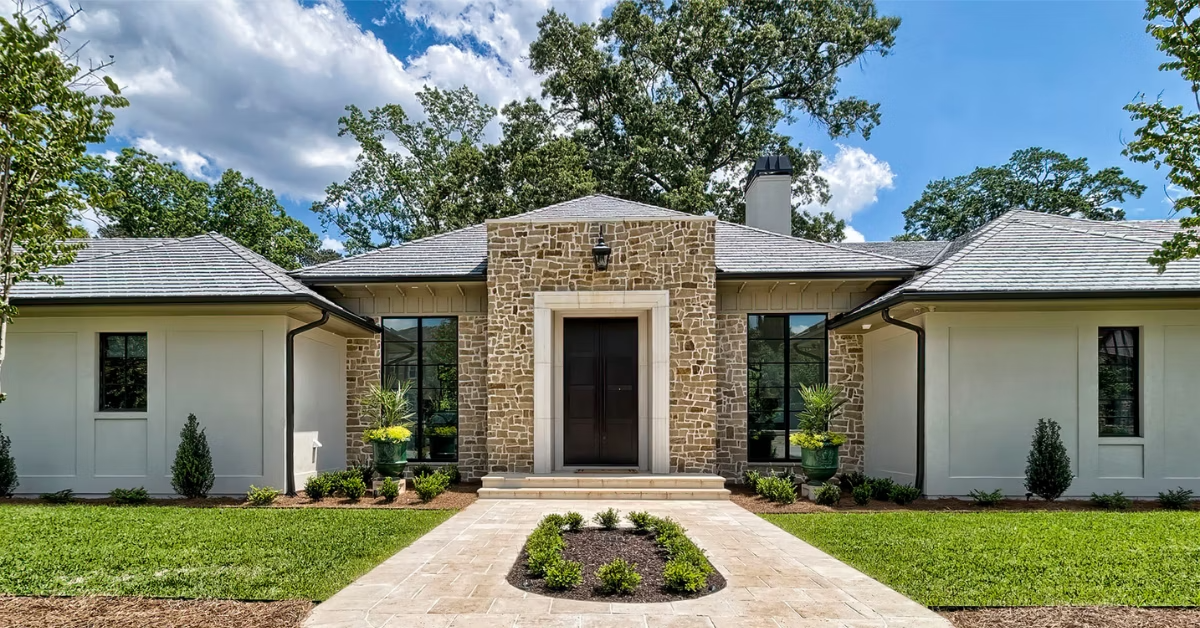 A Mediterranean-style home with low-pitched roof and wide eaves, a stucco veneer, and around the entry way a stone veneer and a sidewalk with landscaping in the center.