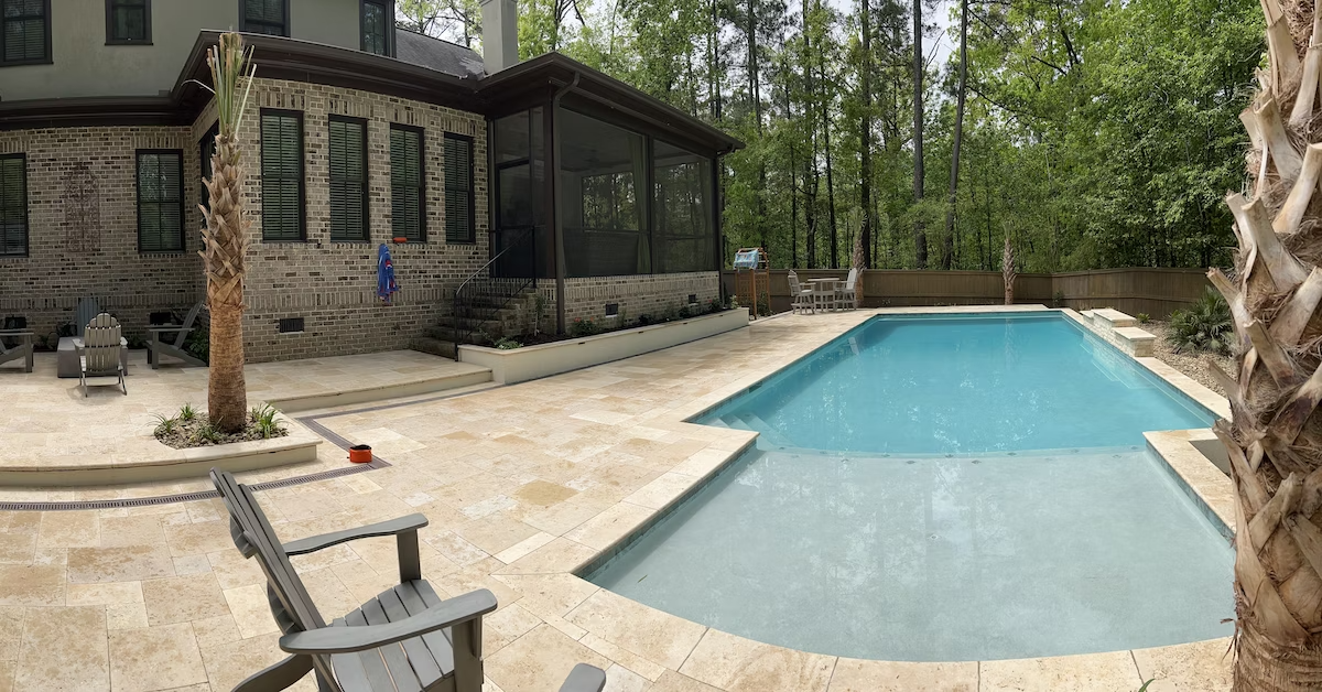 A custom pool with travertine pool deck, large tanning shelf, and waterfall feature behind a brick and stucco Tudor-style home with Palmetto trees and a wood fence.