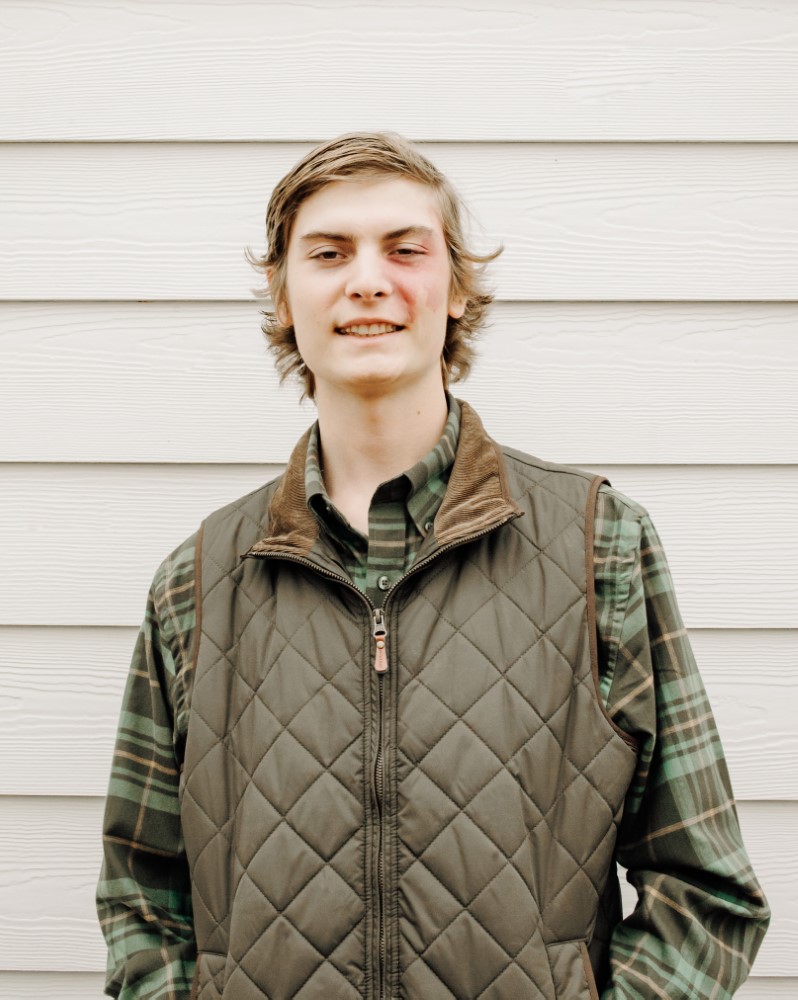Lee Blythe Jr wearing an olive green vest and dark green plaid long-sleeve shirt standing in front of a wall of ivory-colored Hardie plank siding.