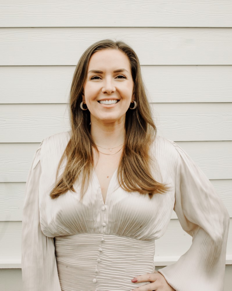 Sarah Bennett wearing a cream-colored blouse standing in front of ivory-colored Hardie siding smiling at the camera.