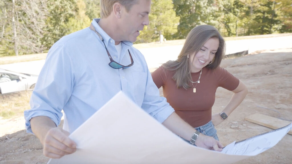 Lee Blythe wearing a long-sleeve blue dress shirt standing on a build site showing Sarah Bennett the house plans.