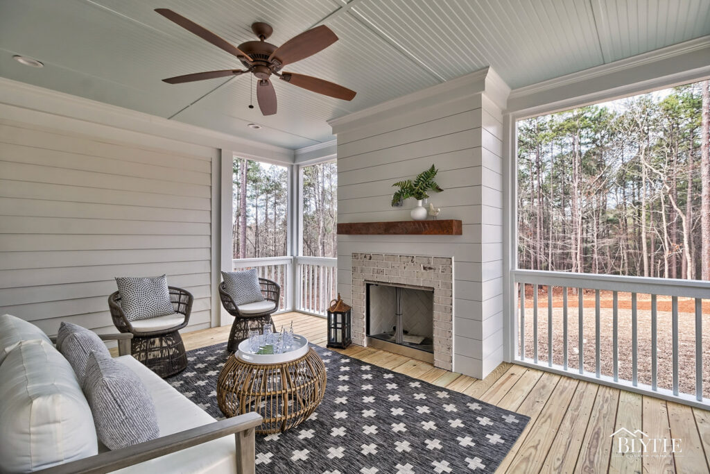 Covered back porch with natural wood decking, painted white beadboard ceiling, and a shiplap wall surrounding the outdoor whitewashed brick fireplace and view of the woods.