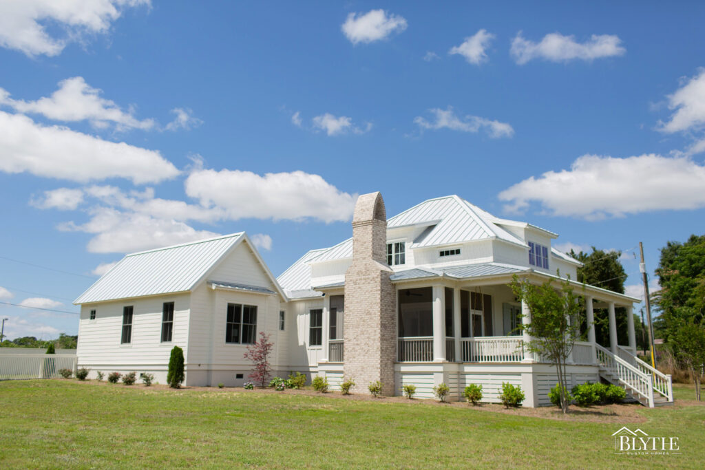 A two-story home with metal roof and shed dormers, a large front porch and screened-in side porch, a brick chimney, and a flat, grassy yard.