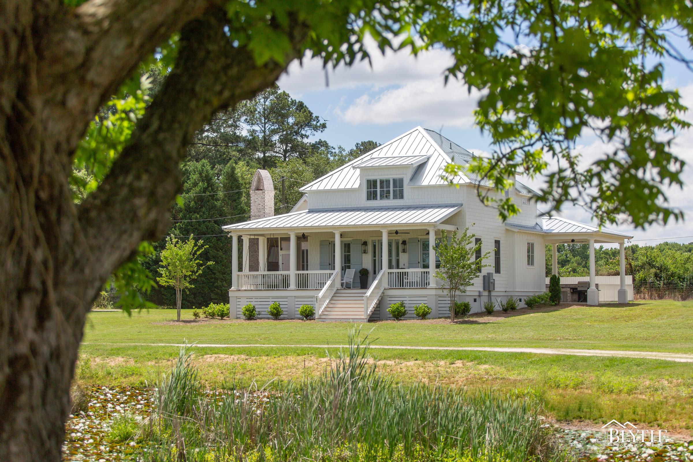 SC Lowcountry Living- Front of home with large front porch, white Hardieplank siding, metal roof, and shed dormer wtih a green leafy tree in the foreground.